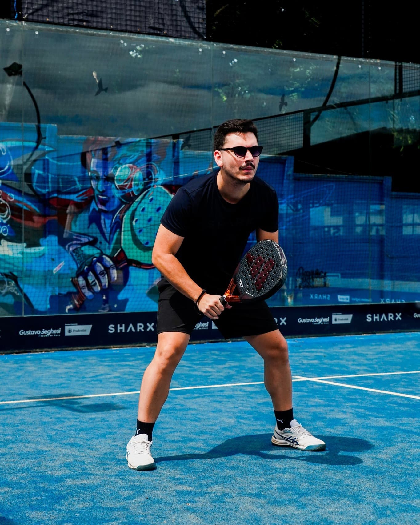 Jogador de padel na quadra azul da Sharx, com o grafite atrás do vidro ao fundo