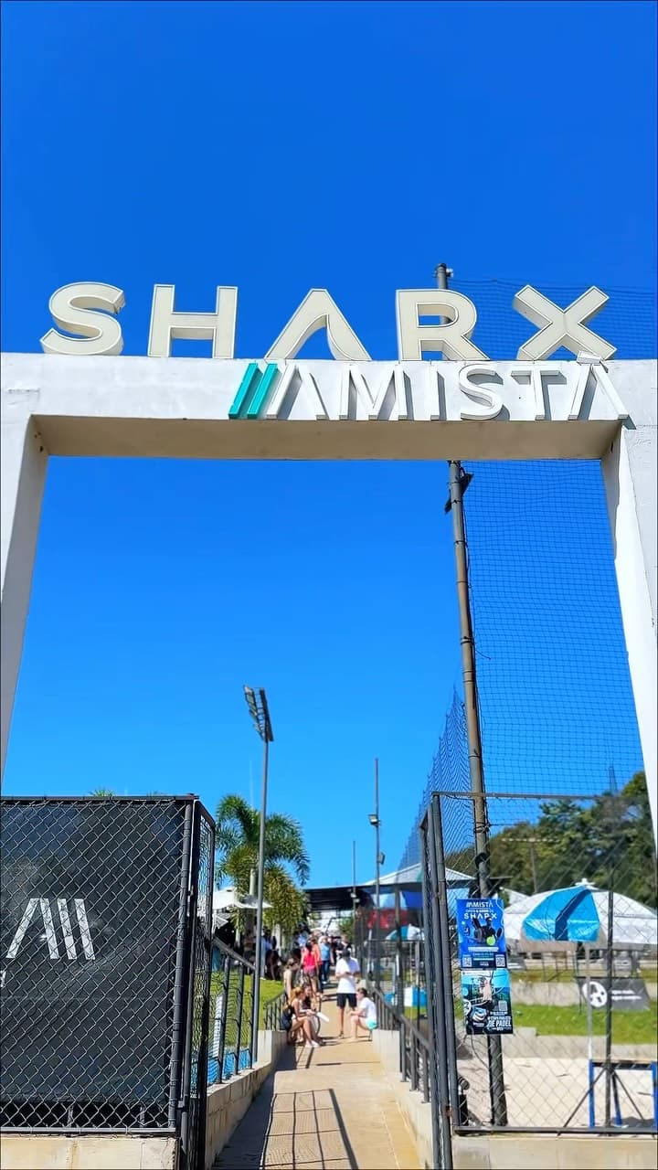 Portal de entrada da arena com o letreiro SHARX Amistà contra o céu azul e o caminho até as quadras