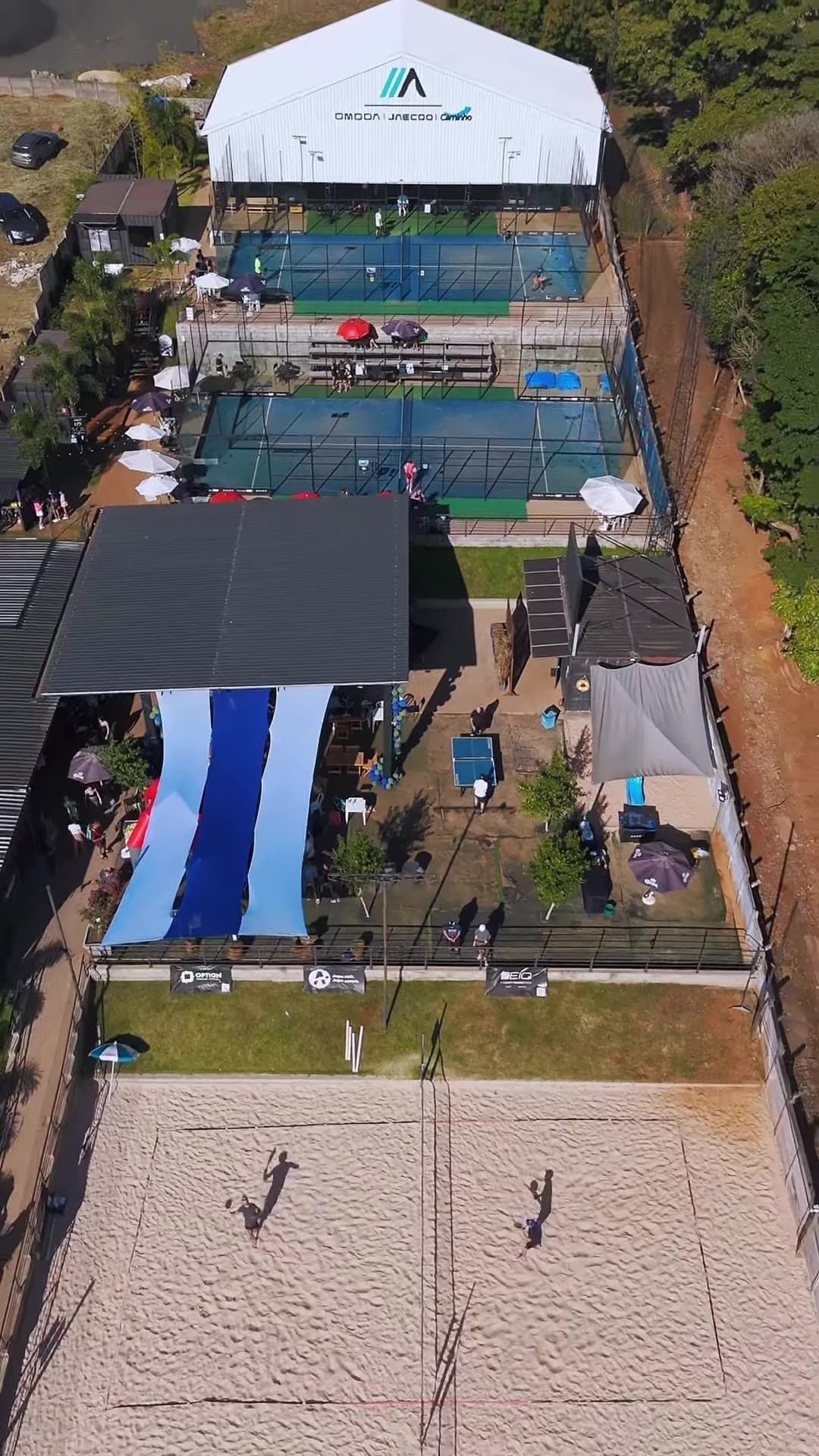 A arena vista de cima: a tenda do padel coberto, as quadras azuis, o deck e as quadras de areia
